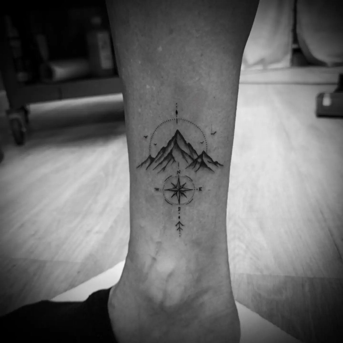 Ankle tattoo featuring mountain peaks and a compass rose in black ink.