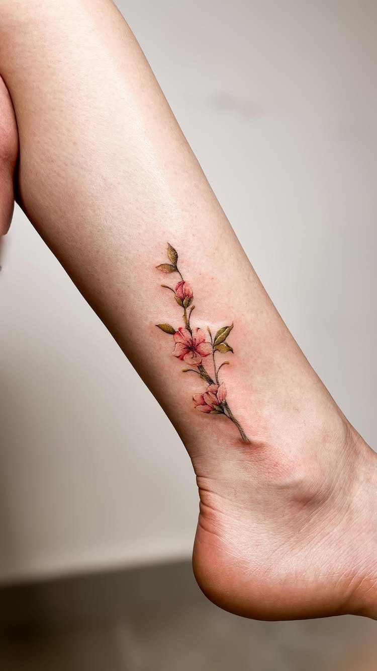 Flowering branch tattoo with pink blossoms and green leaves on a person's ankle