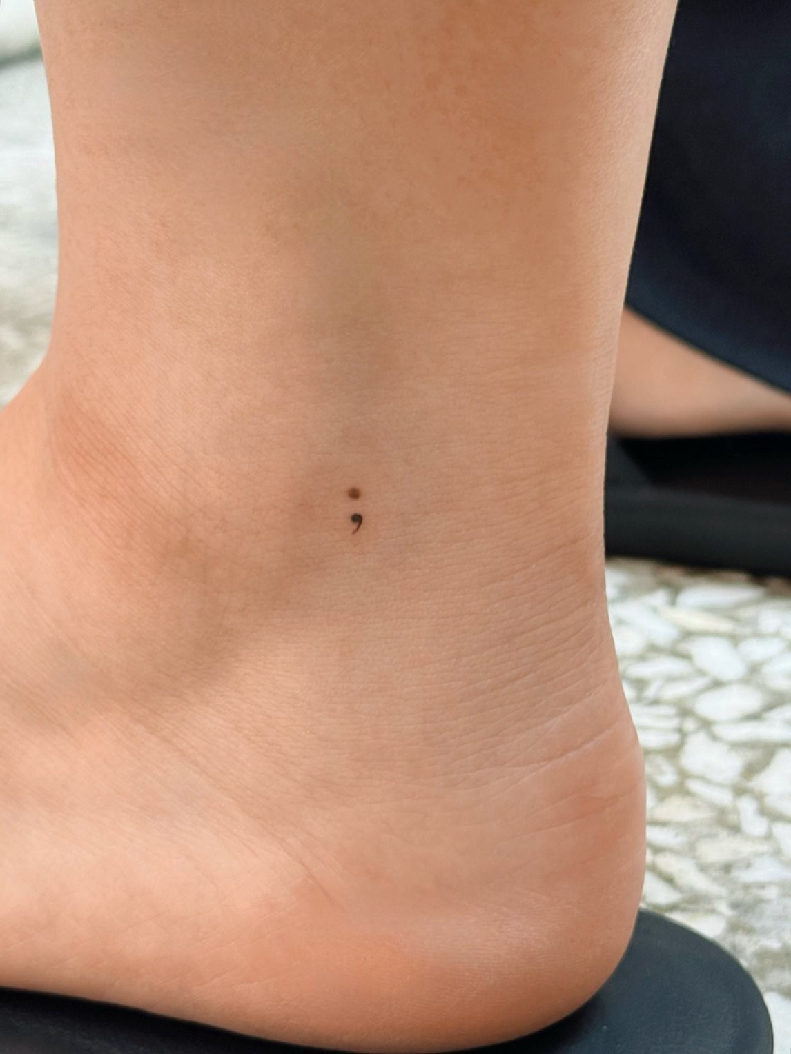 Small black semicolon tattoo on the inner ankle of a person's foot.