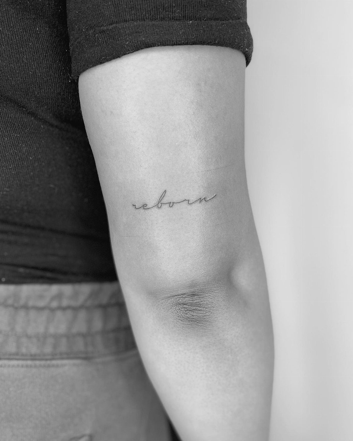 Cursive reborn tattoo on the back of a person's upper arm.