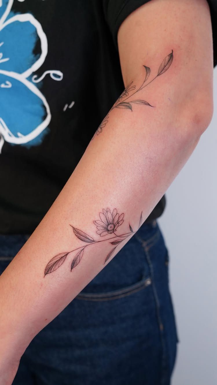 Small daisy tattoo with leaves on the back of a person's forearm.