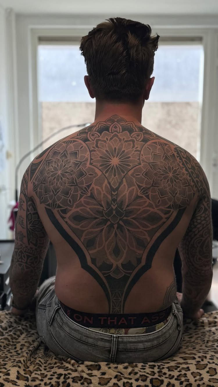 Man's back featuring a large, symmetrical black ink mandala tattoo with intricate patterns.