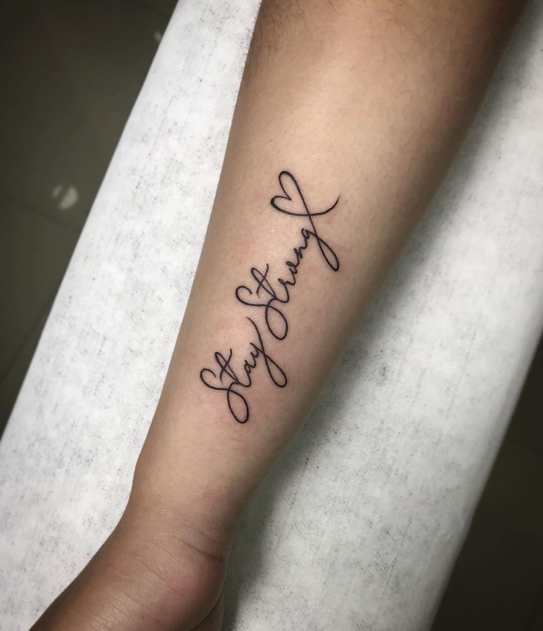 Small black ink cursive tattoo reading Stay Strong on a person's forearm