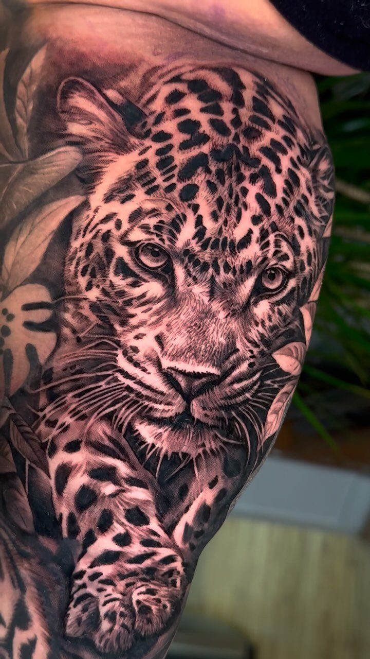 Leopard realism tattoo on a woman's bicep with detailed shading and tropical leaves.