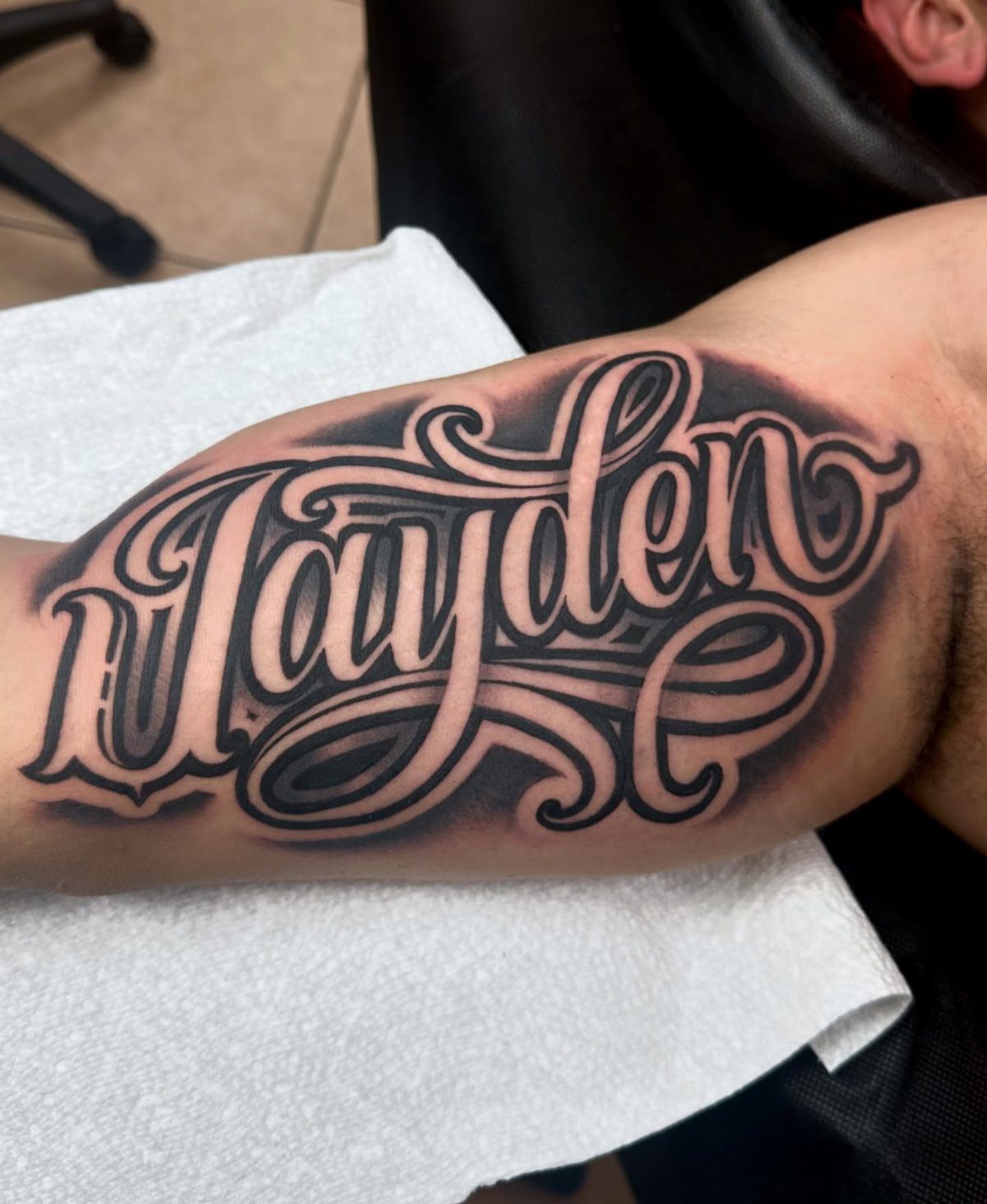 Stylized cursive tattoo of the name Jayden on a man's bicep arm.