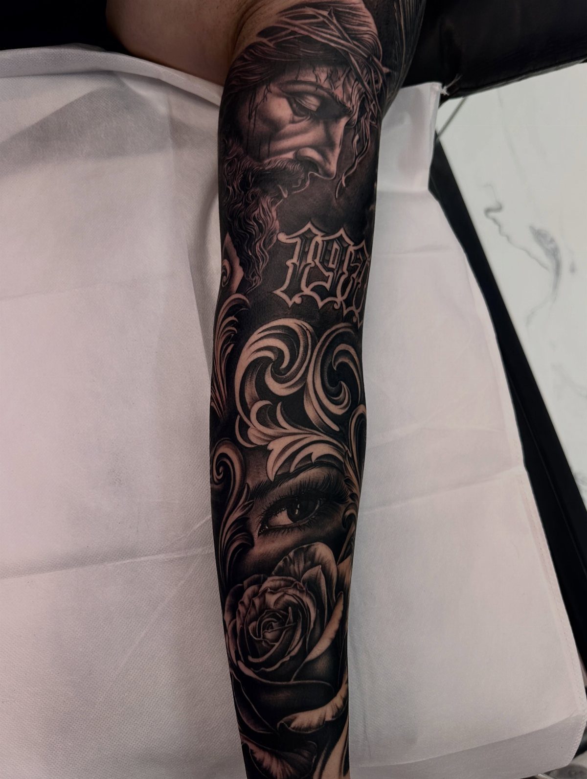 Realism tattoo on an arm featuring Jesus, a rose, and an eye in black ink.