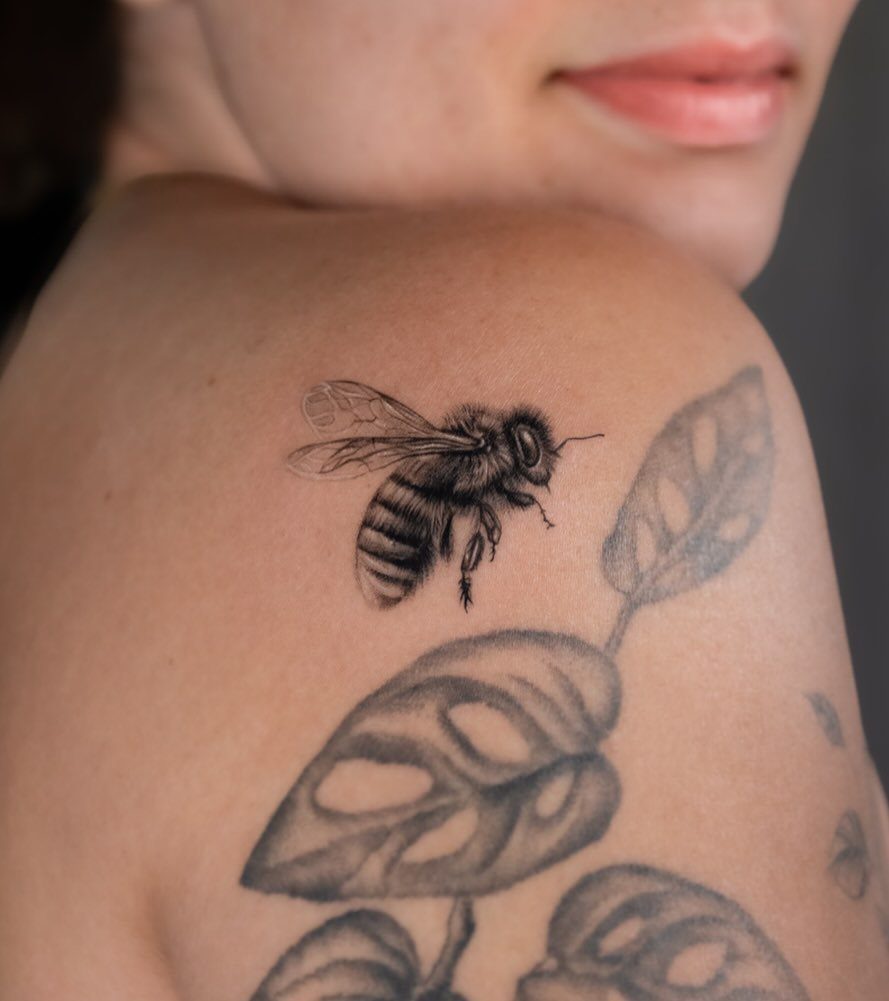 Honeybee tattoo on a woman's shoulder with accompanying botanical leaf designs.