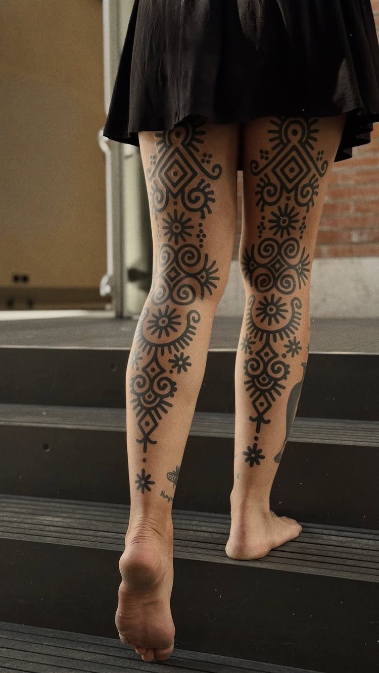 Woman's legs featuring bold black tribal tattoos with geometric patterns on dark stairs.