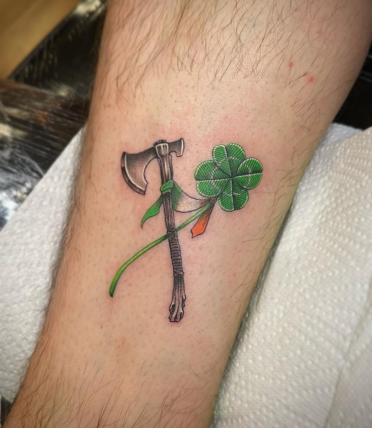 Axe tattoo with a green four-leaf clover on a man's hairy calf.