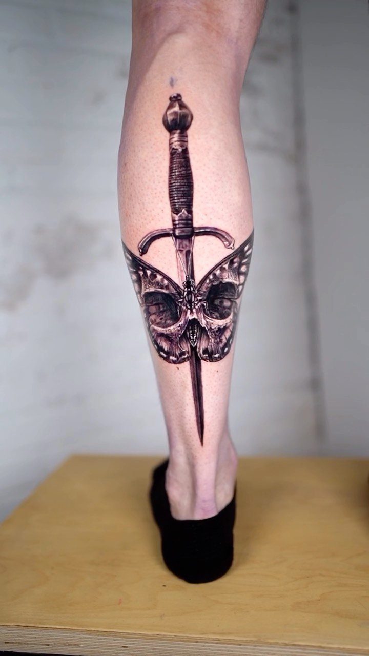 Dagger tattoo piercing a skull with butterfly wings on a man's calf