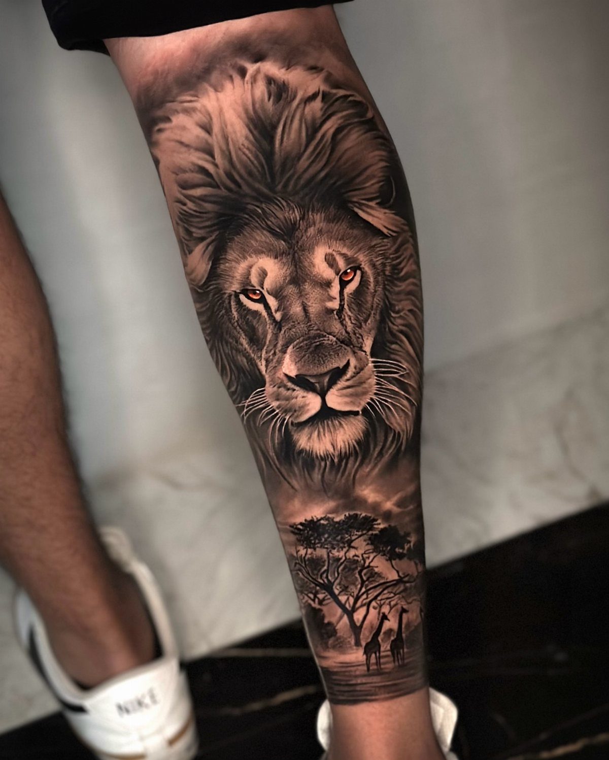 Lion tattoo on a man's calf with red eyes and savanna landscape below.