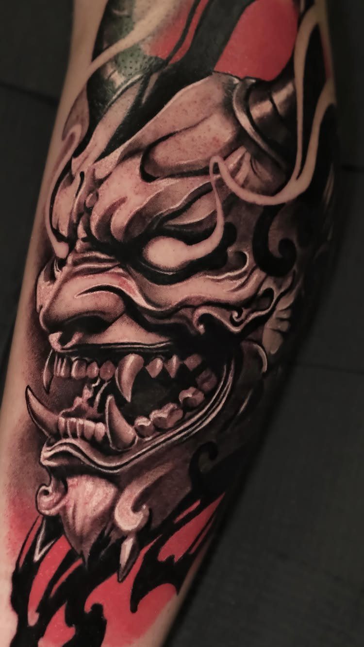 Oni mask tattoo on a calf with grey shading and red accents.