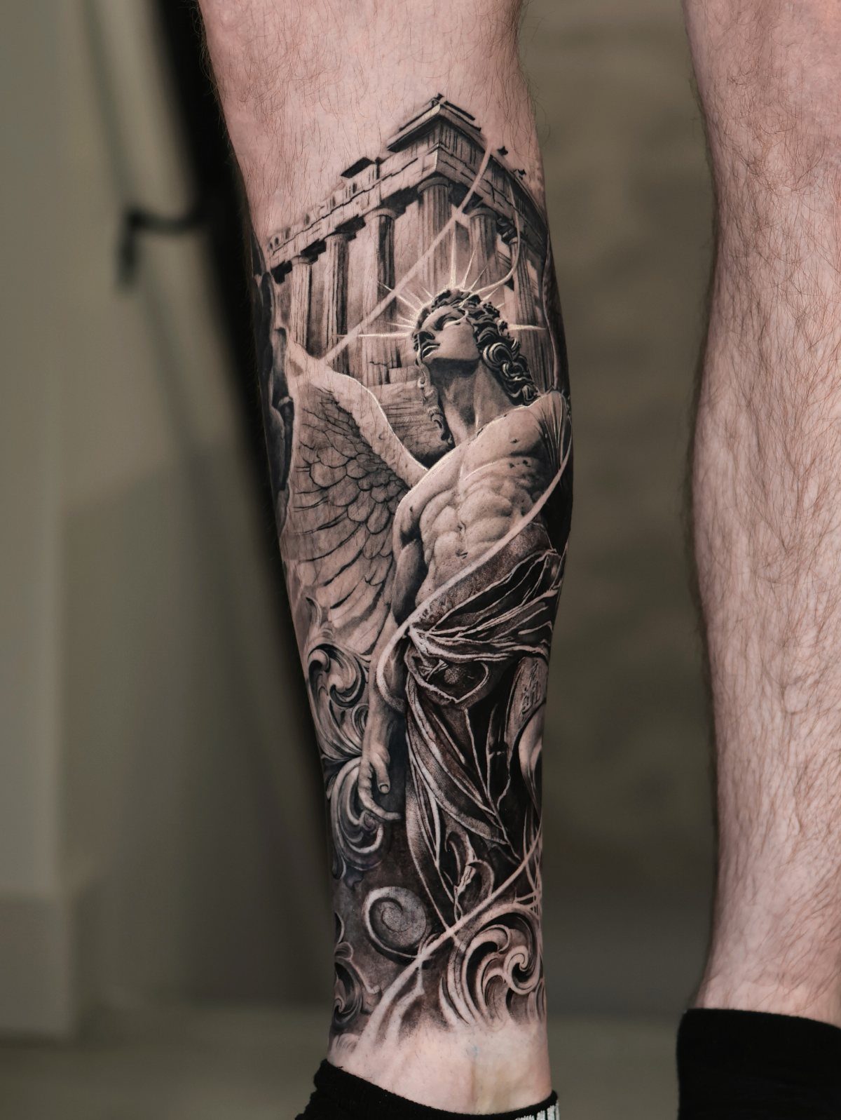 Realistic black and grey tattoo of a winged statue on a man's calf.