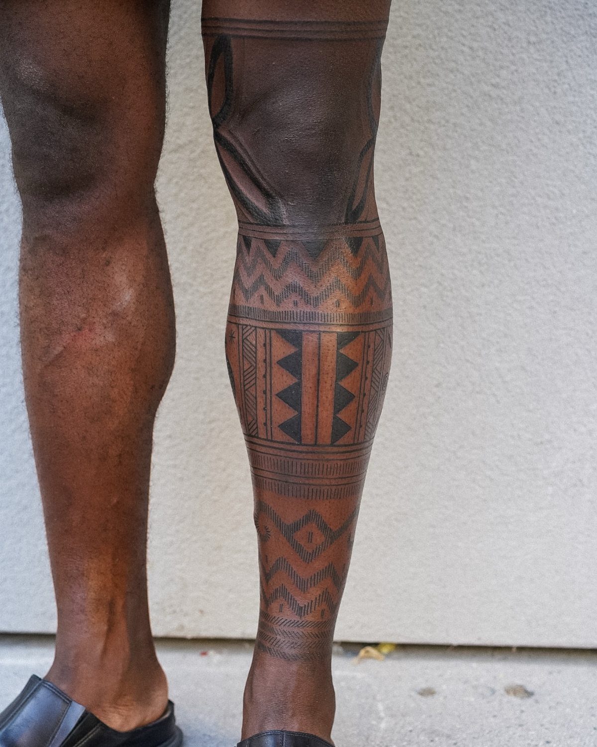 Man's calf with a bold black geometric tribal tattoo against a white wall.