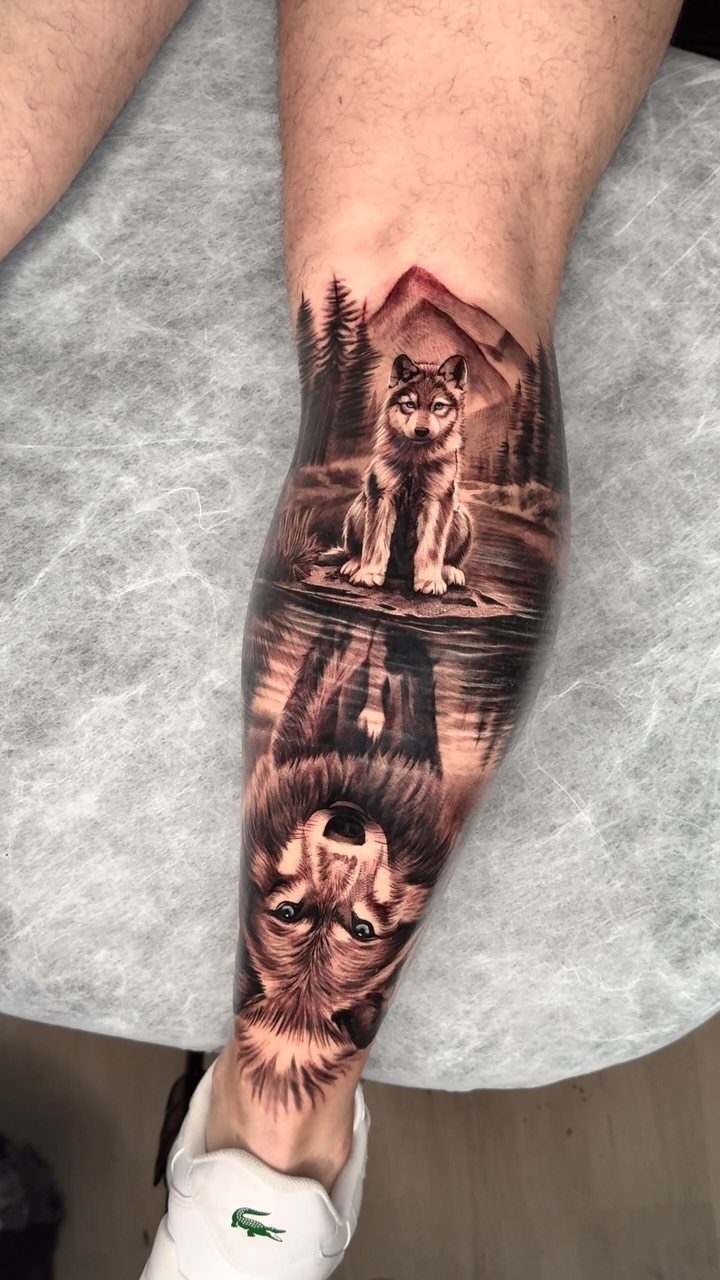 Wolf tattoo on a man's calf with a mountain landscape and water reflection.