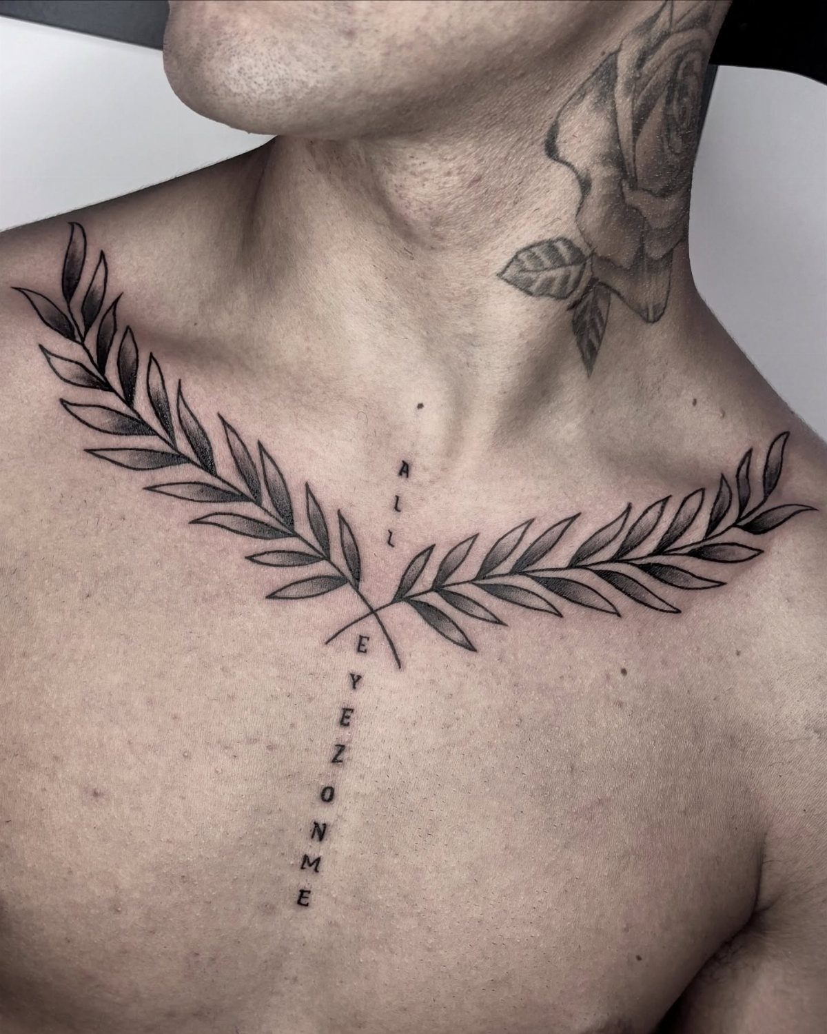 Laurel wreath tattoos along the collarbones with vertical text on the center chest.