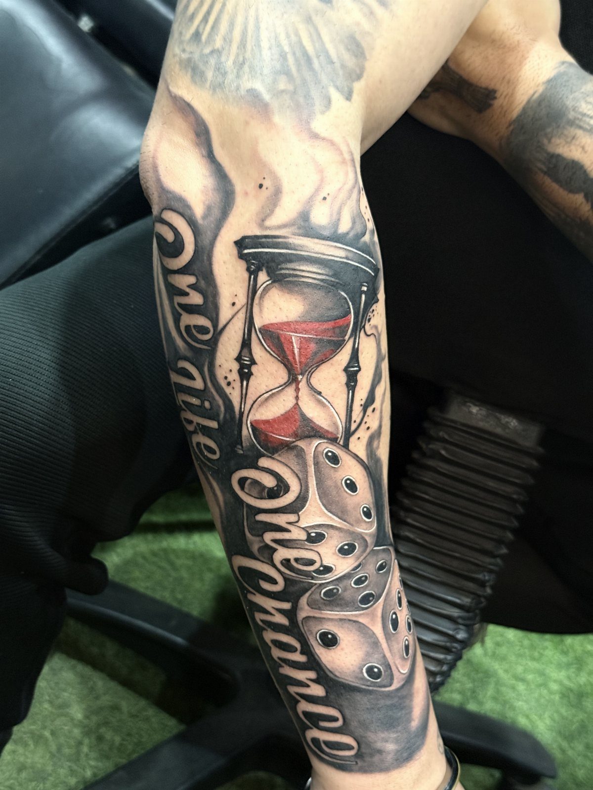 Forearm tattoo featuring an hourglass with red sand and dice amidst grey clouds.