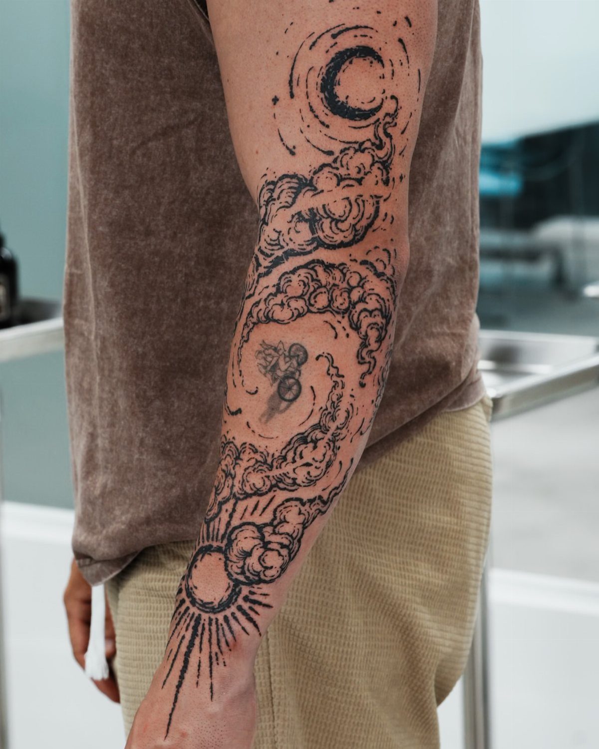 Forearm tattoo featuring swirling black ink clouds with a crescent moon and sun.