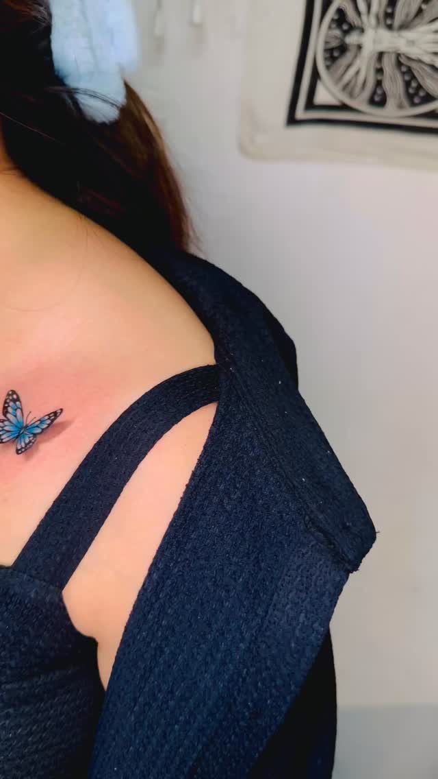 Small blue butterfly tattoo on a person's collarbone area wearing black clothing.