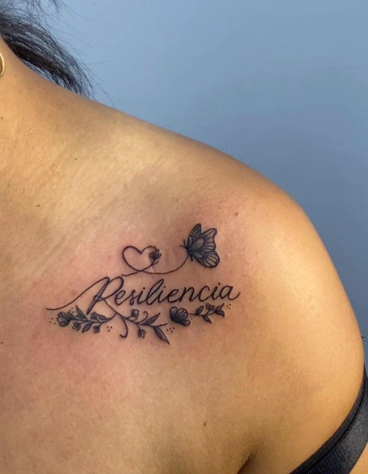 Cursive tattoo of the word Resiliencia with a butterfly and flowers on a collarbone.