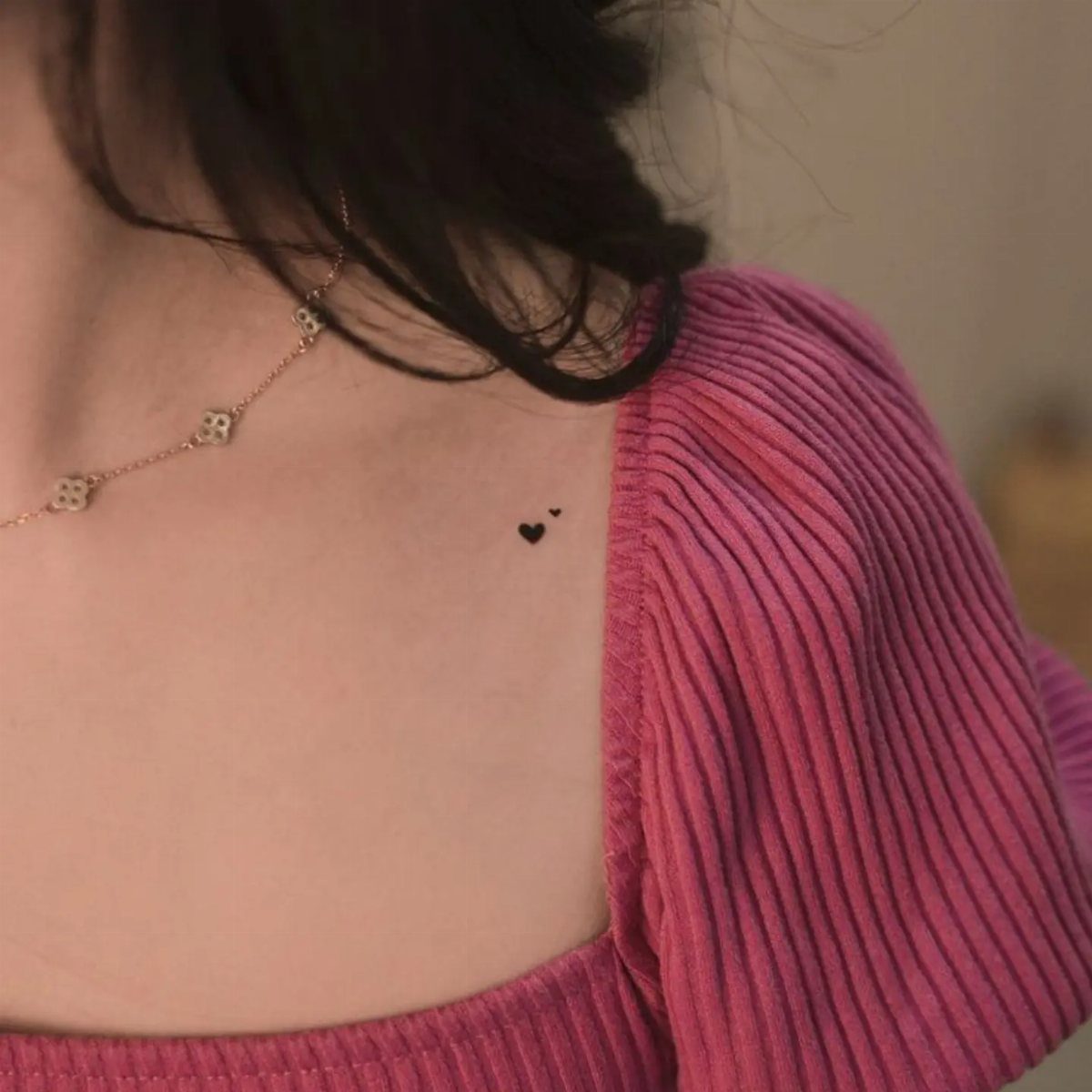 Two tiny black heart tattoos on a person's collarbone wearing a pink top.