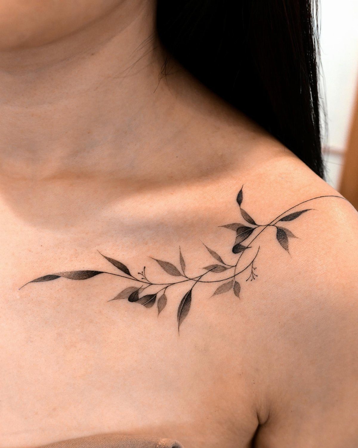 Black and grey vine tattoo with delicate leaves on a woman's collarbone.