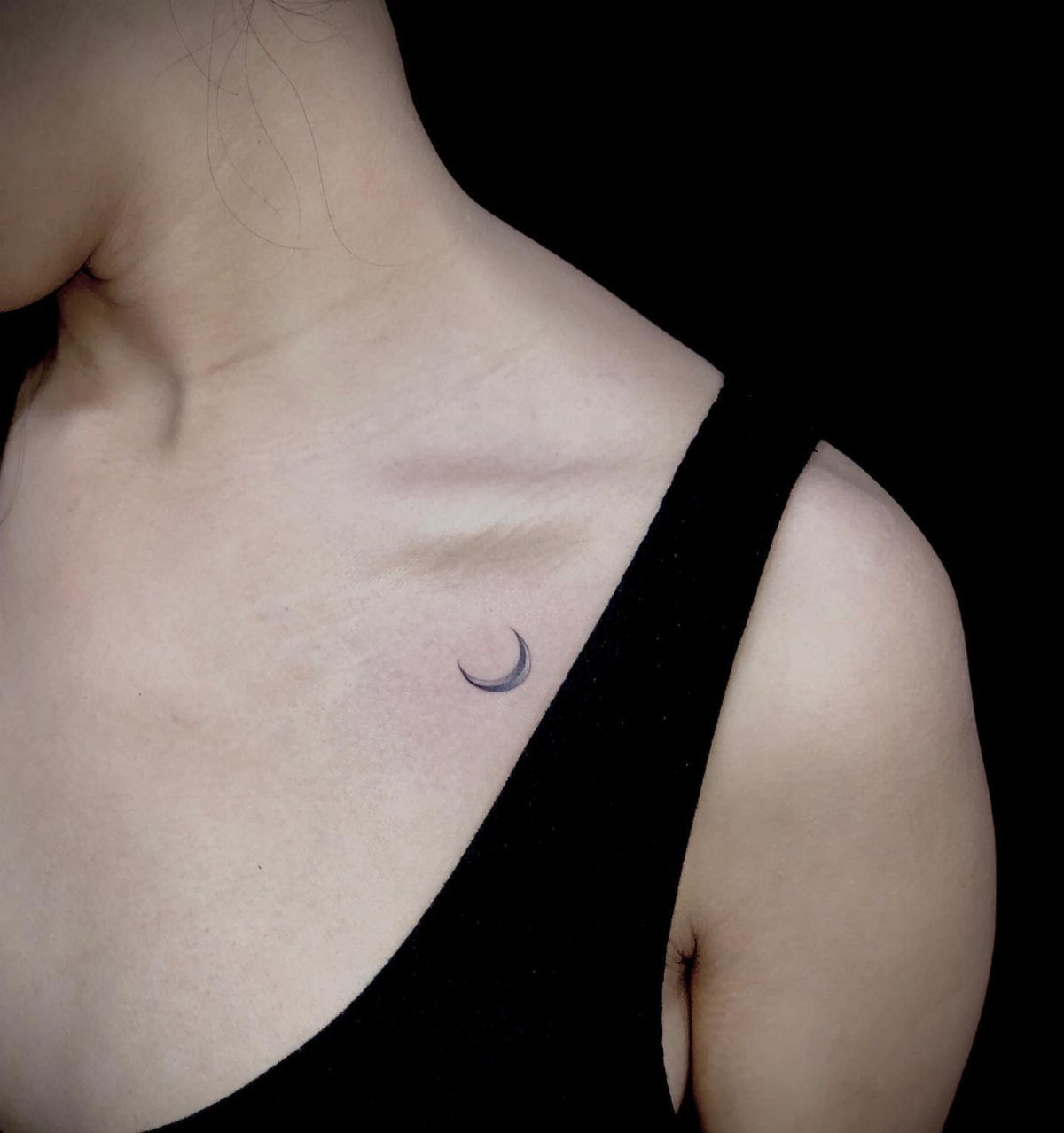 Small black crescent moon tattoo on a woman's collarbone area.