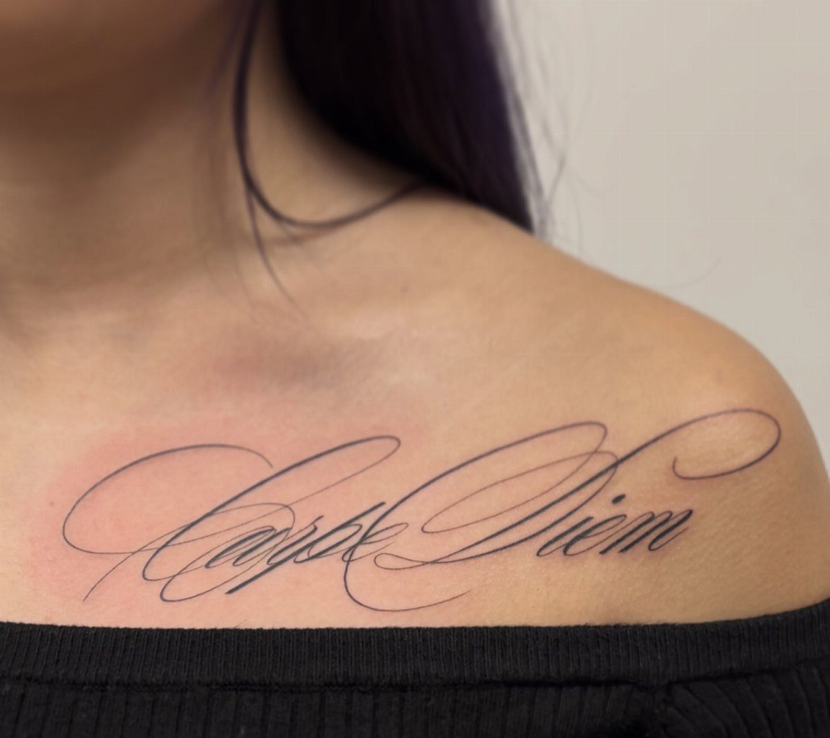 Cursive Carpe Diem tattoo on a woman's collarbone with a black off-shoulder top.