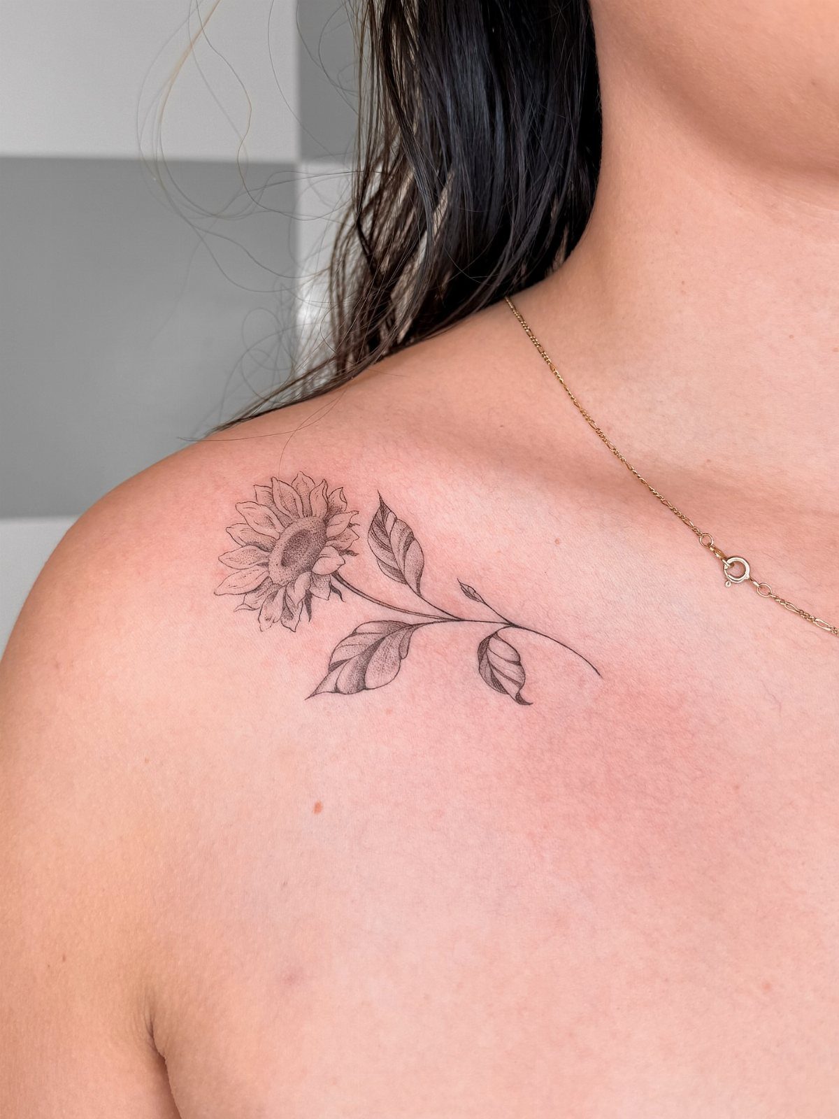 Sunflower line-art tattoo on a woman's collarbone and shoulder with a gold necklace.