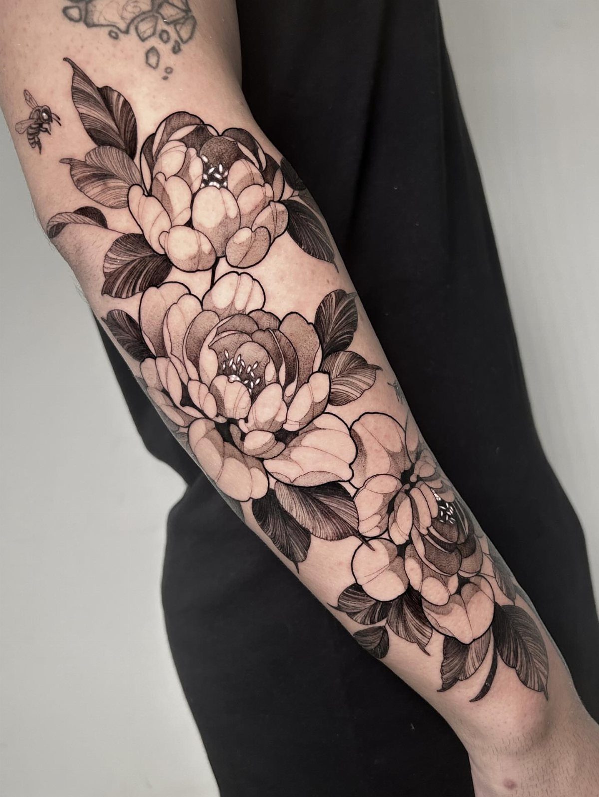 Blackwork peony flower tattoo with leaves and a bee on a forearm