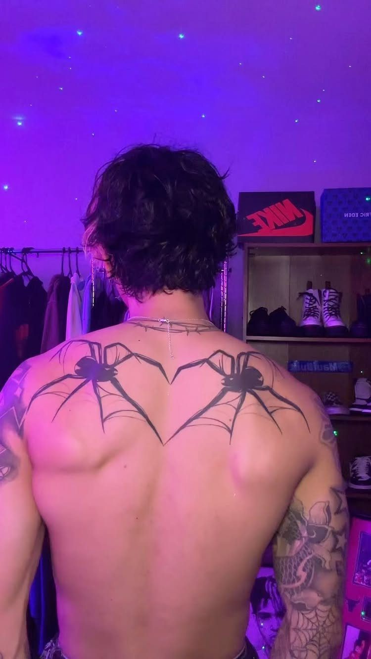 Man's back with a large black spider web heart tattoo under purple lighting
