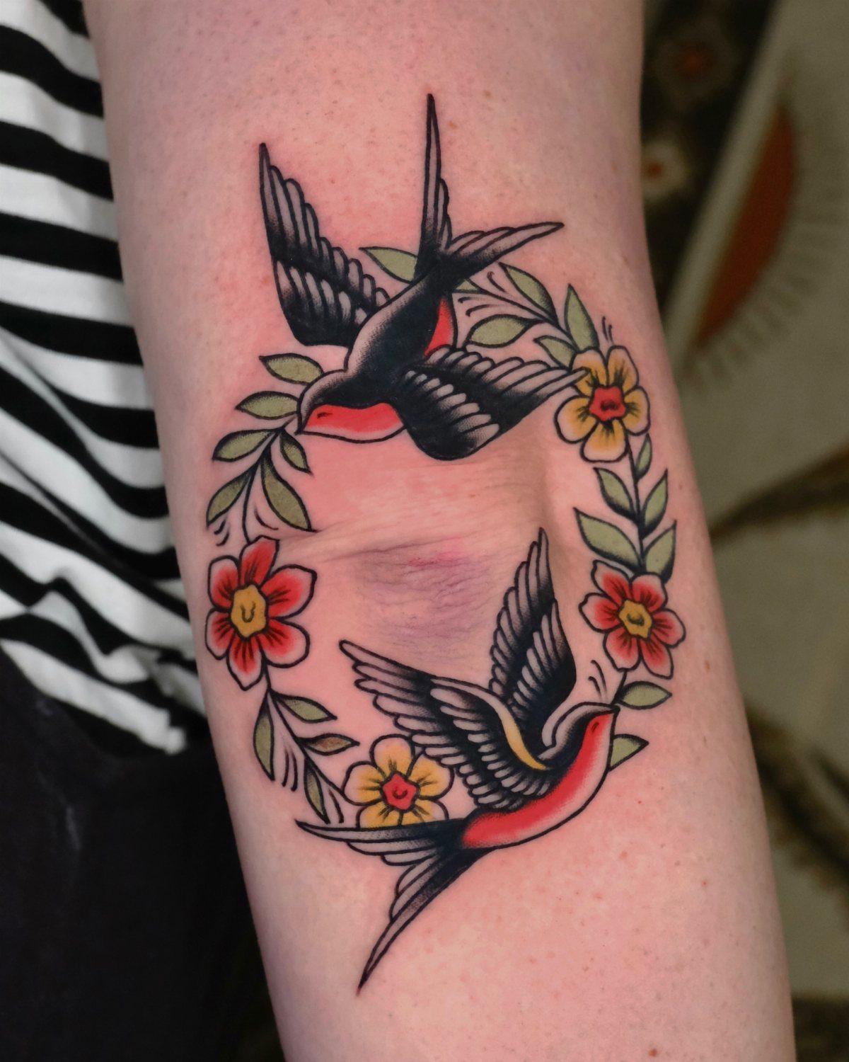 Two swallows and colorful flowers tattooed in American traditional style on an elbow.