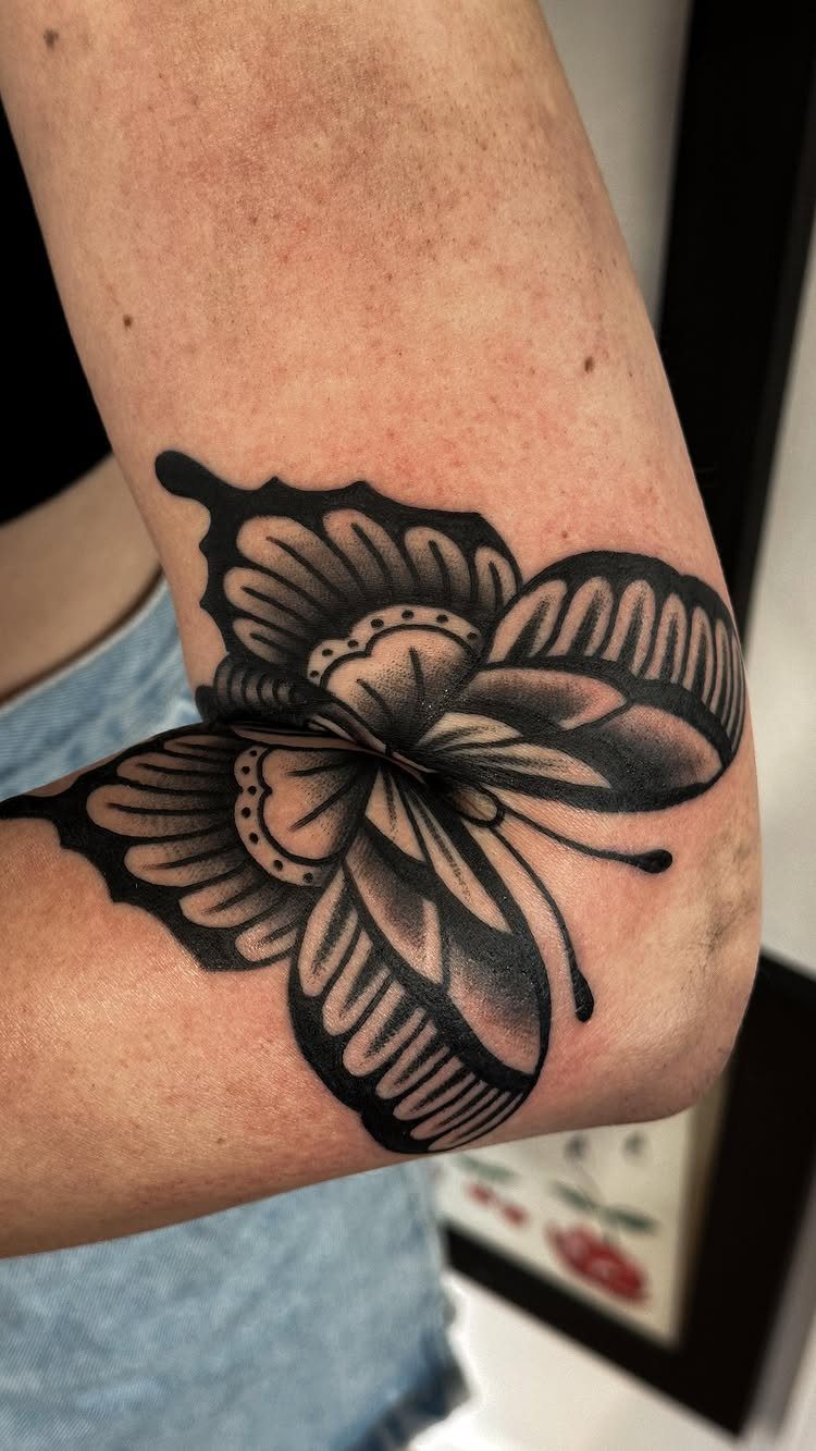Symmetrical butterfly tattoo in black and grey ink centered on a person's elbow.