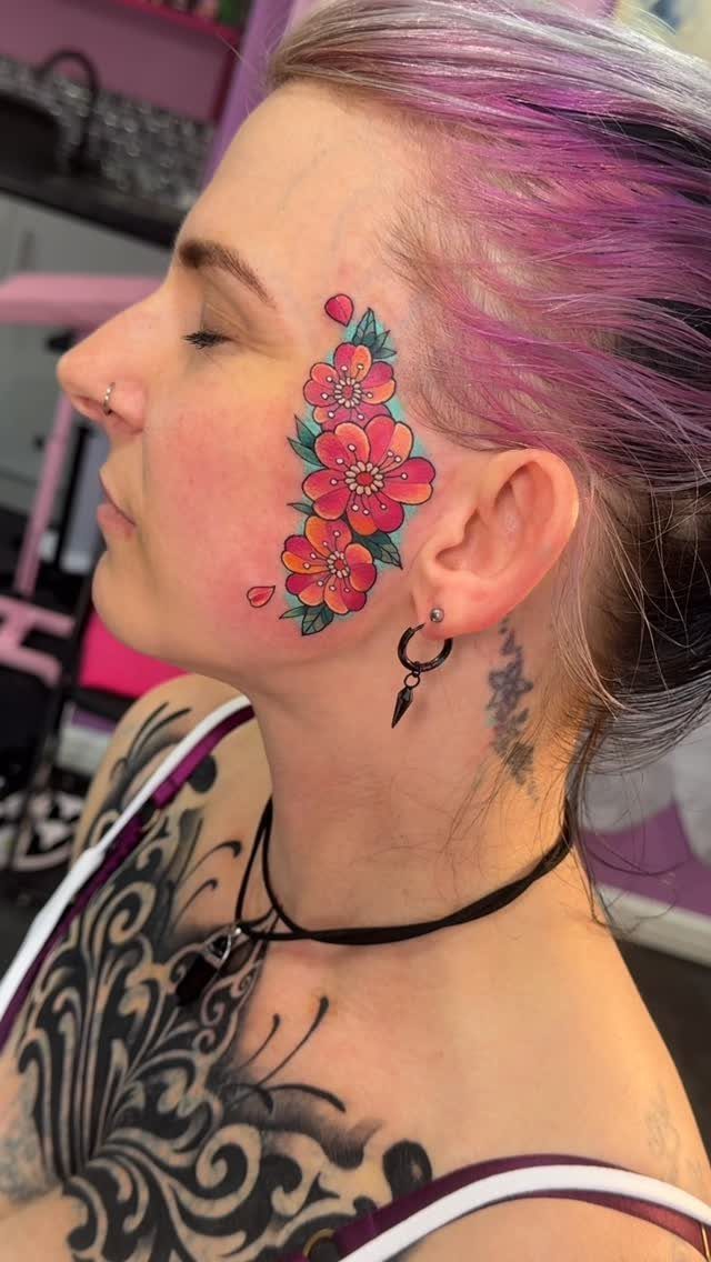 Person with purple hair and a colorful floral tattoo on their cheek.