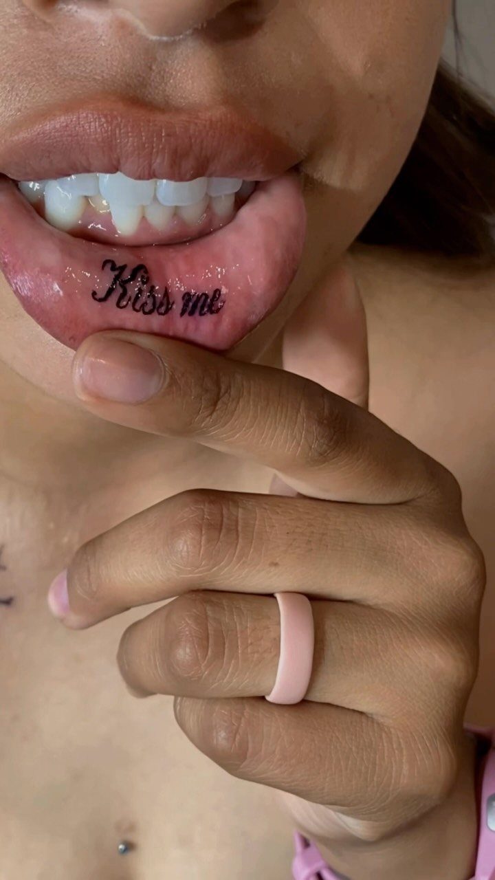 Inner lip tattoo with the words Kiss me in black cursive script.