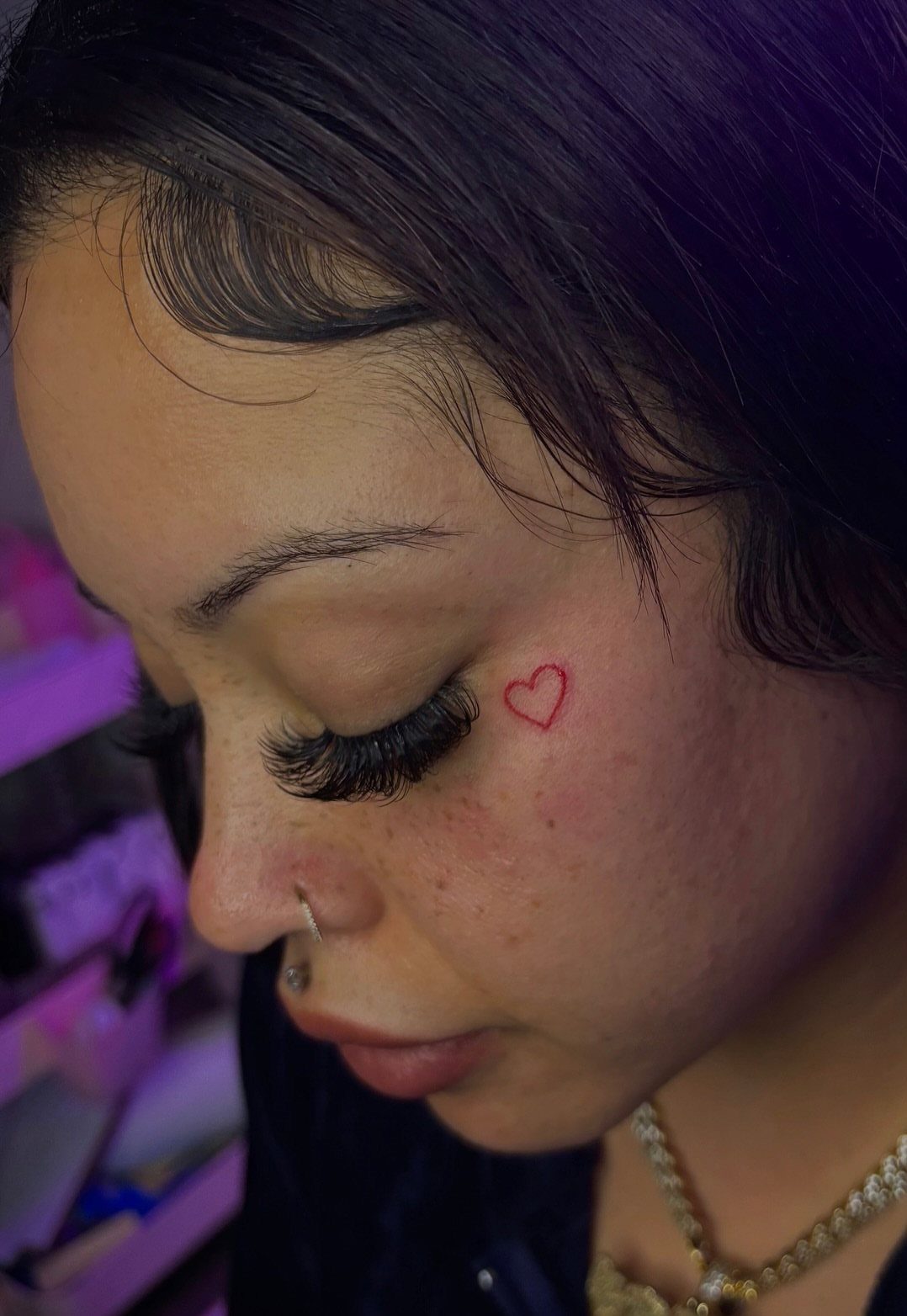 Small red heart tattoo on the cheek of a person with long eyelashes.