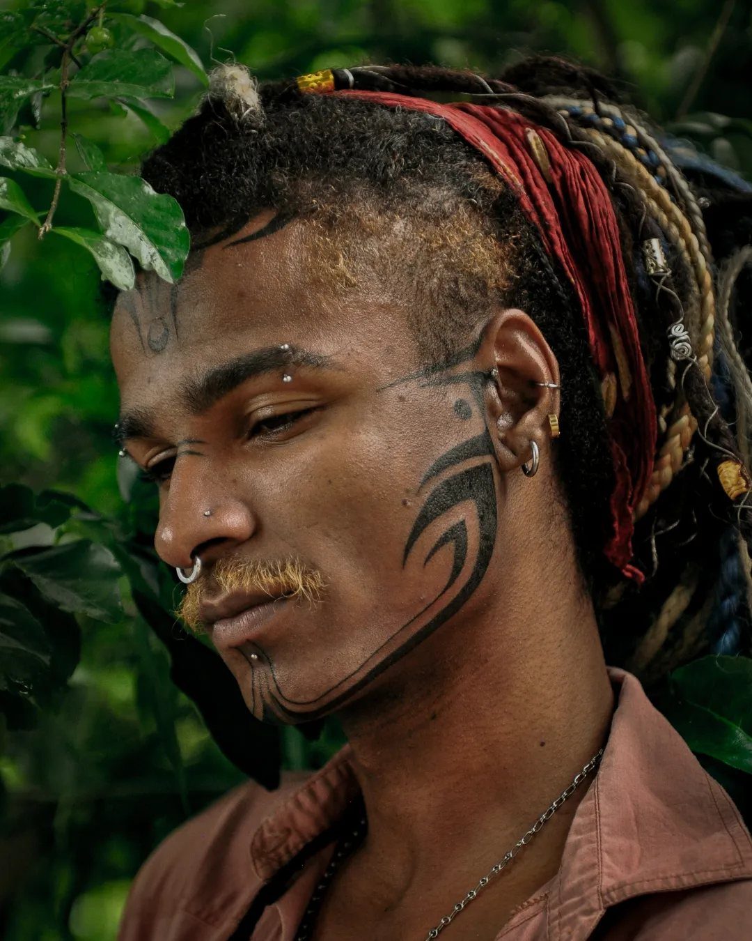 Man with bold black tribal tattoos on his face and jawline among green leaves