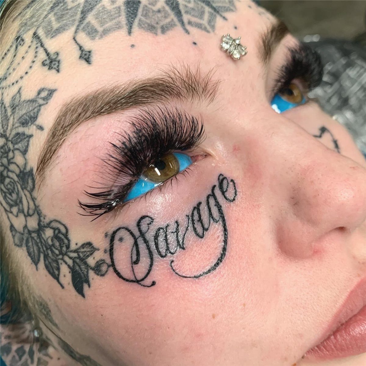 Person with 'Savage' script tattoo under eye and floral tattoos on temple.