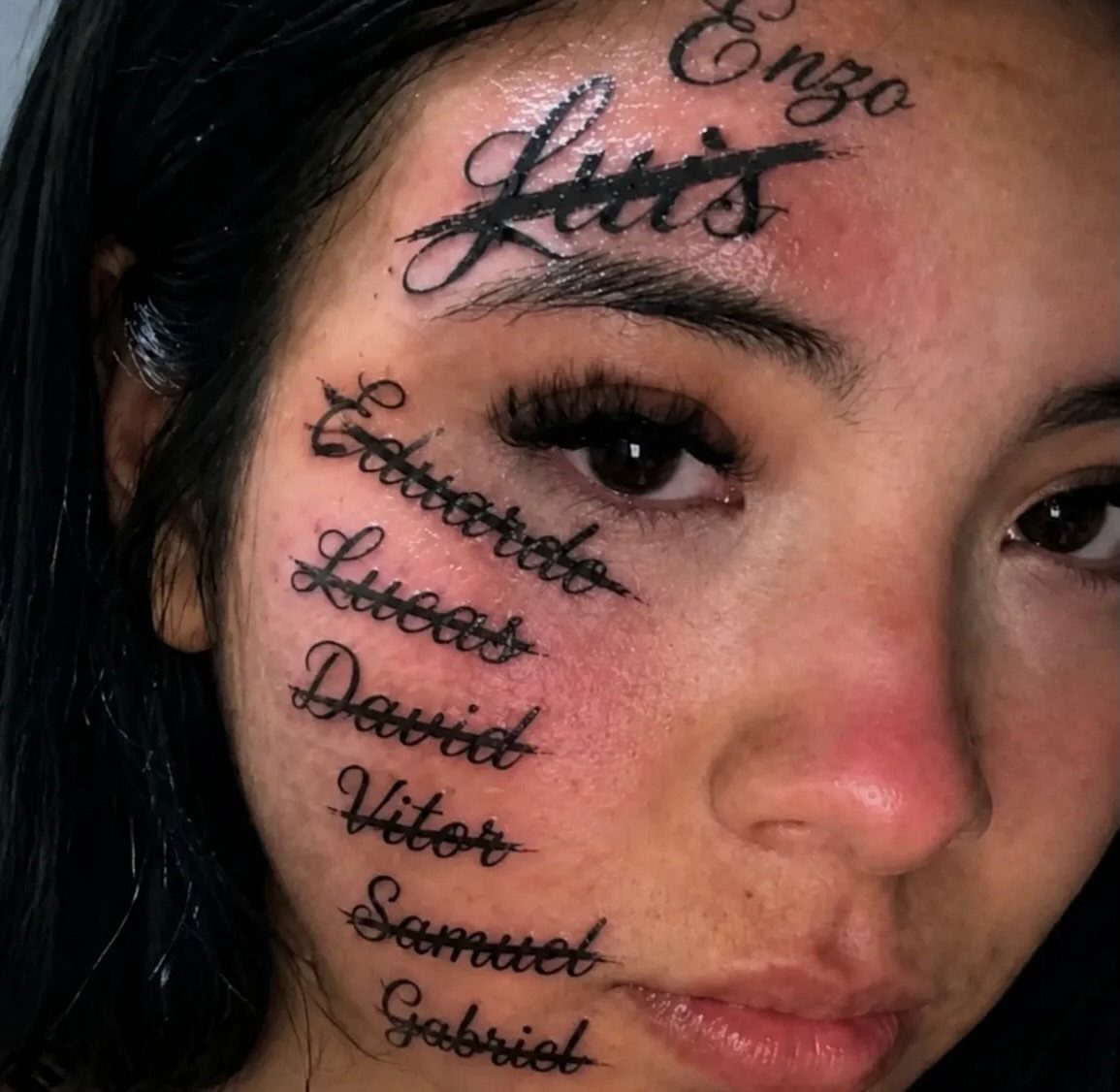 Woman with multiple black ink name tattoos on her forehead and right cheek.