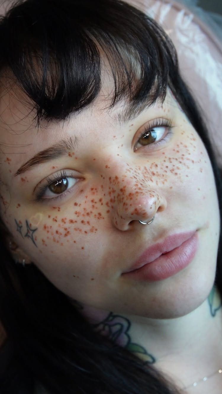 Woman with brown eyes and tattooed freckles across her nose and cheeks.