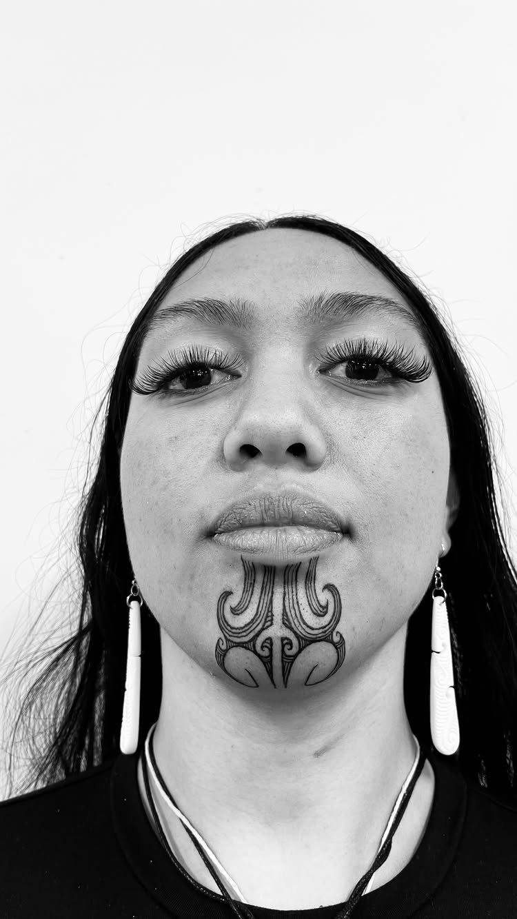 Woman with a traditional Maori Moko Kauae chin tattoo and long white earrings.