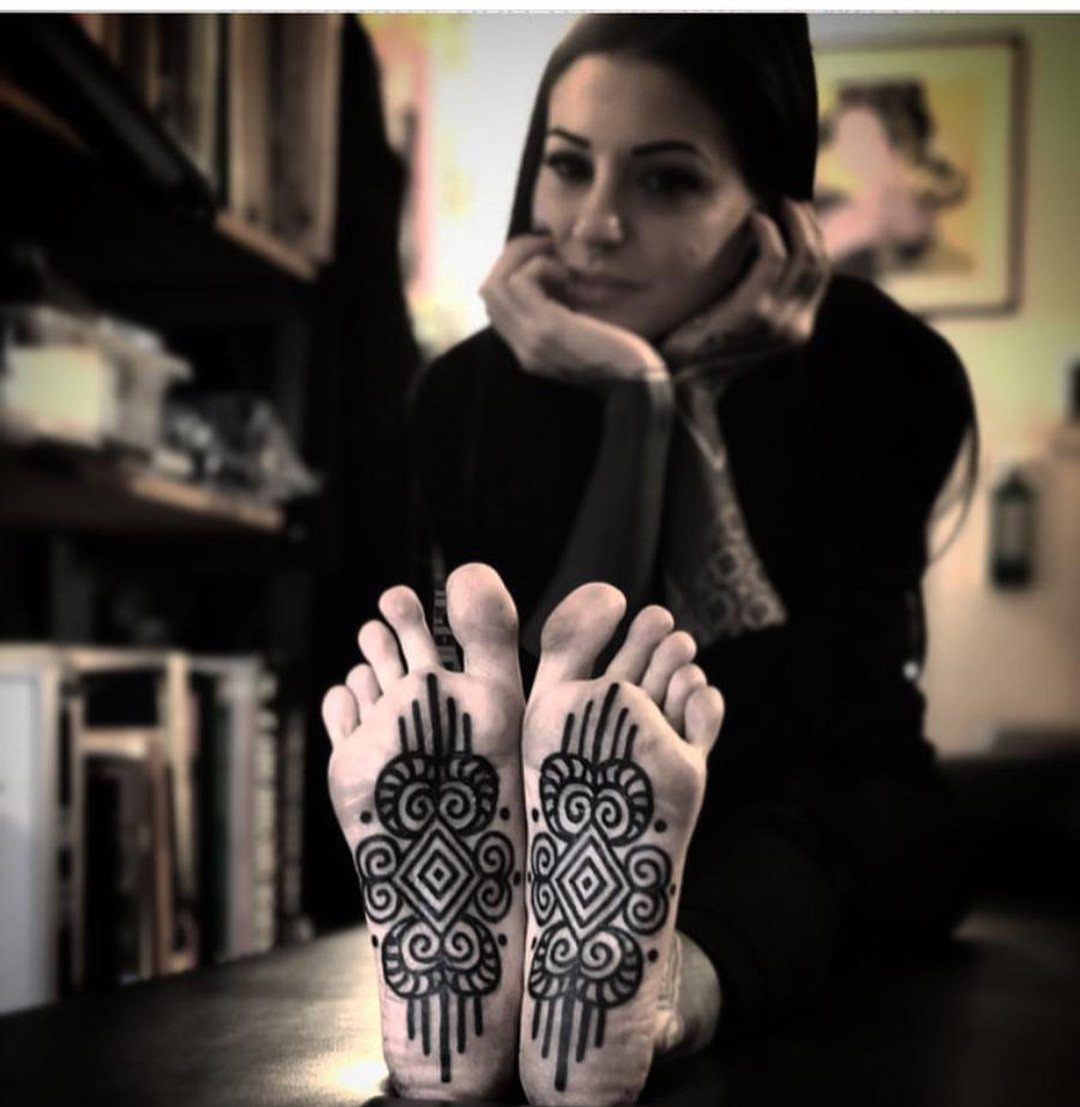 Symmetrical black ornamental tattoos on the soles of a woman's feet.