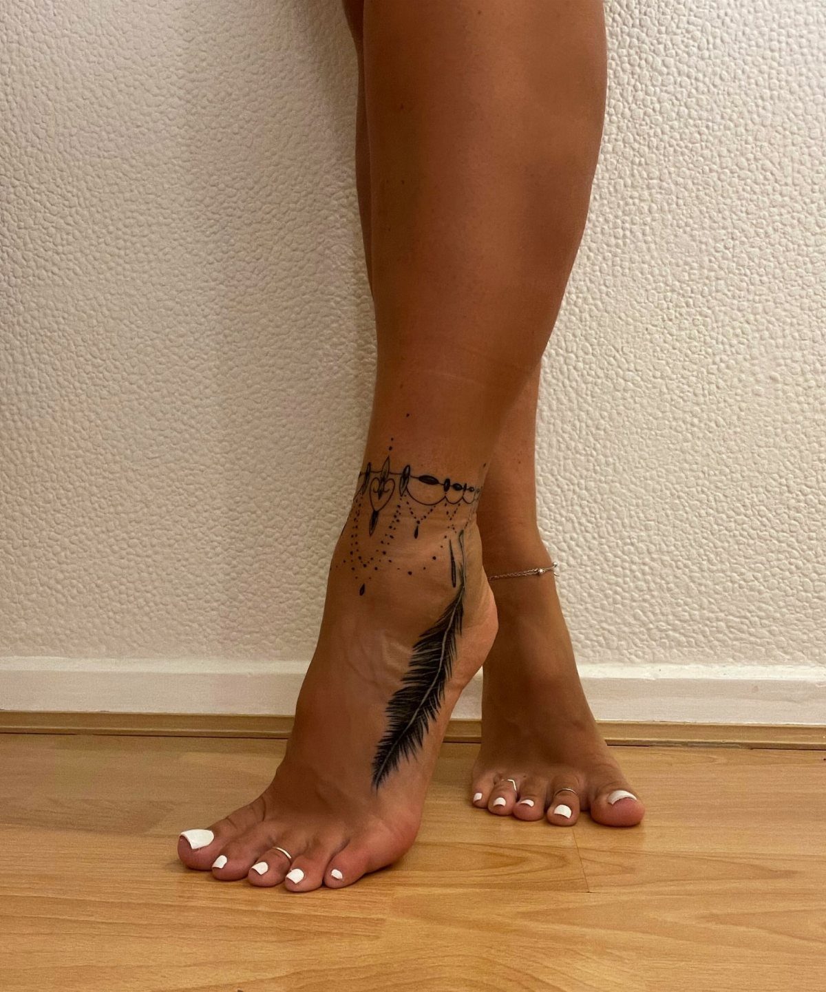 Woman's foot and ankle featuring a black ink feather tattoo and ornamental anklet.