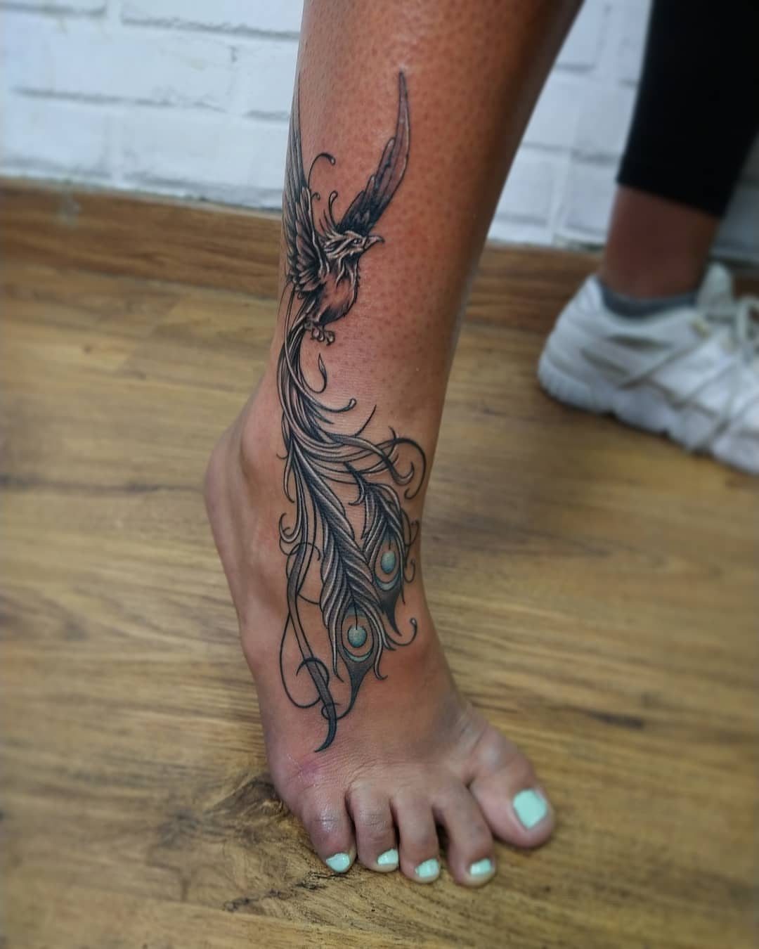 Phoenix tattoo on a woman's foot and ankle with detailed flowing tail feathers.