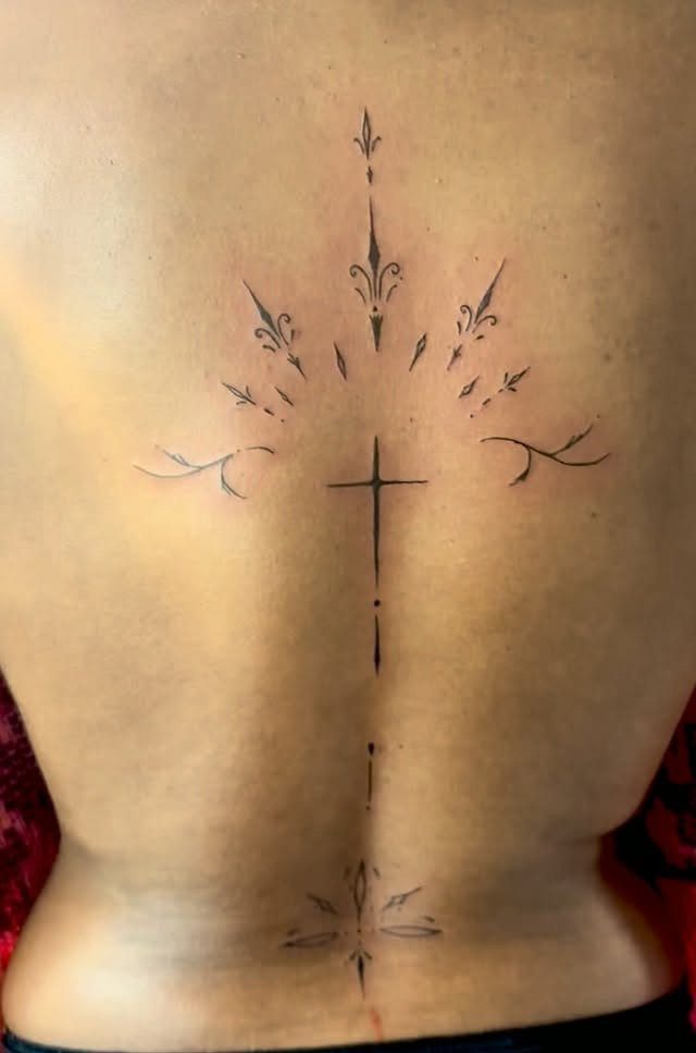 Black ink cross tattoo with ornamental flourishes centered on a person's back.