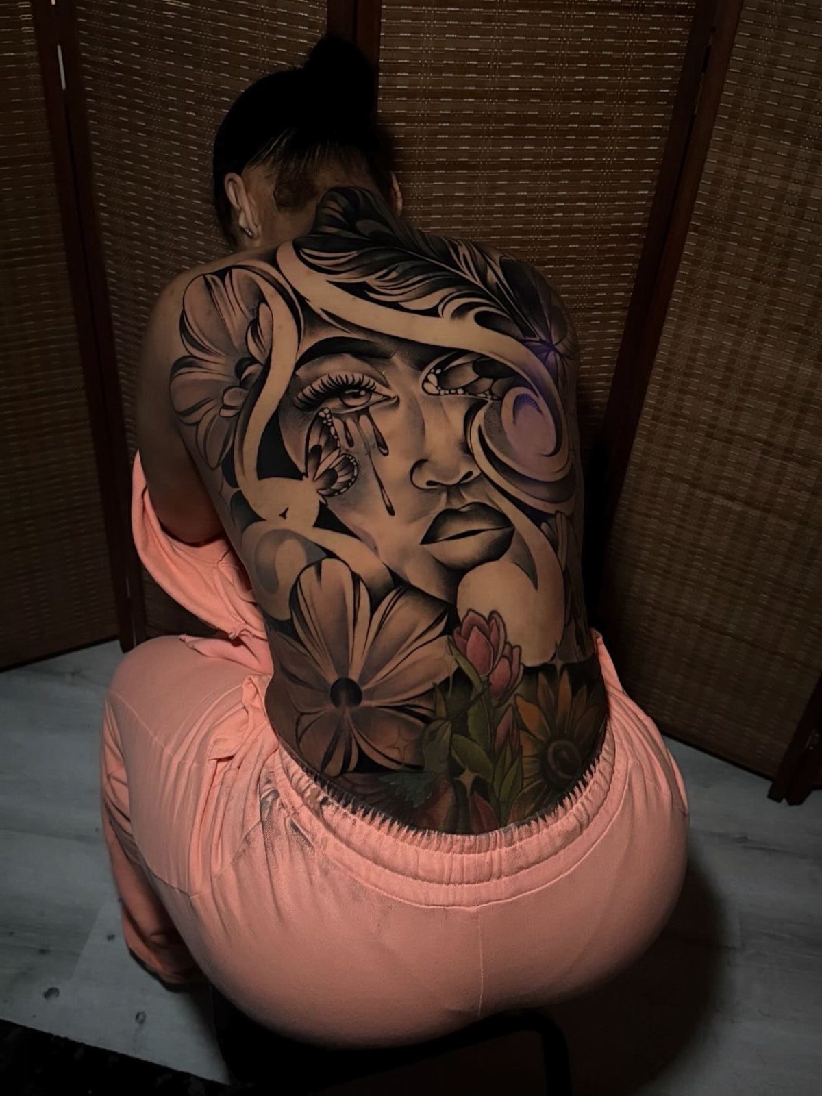 Woman's back featuring a hyperrealistic black and gray tattoo of a face and flowers.