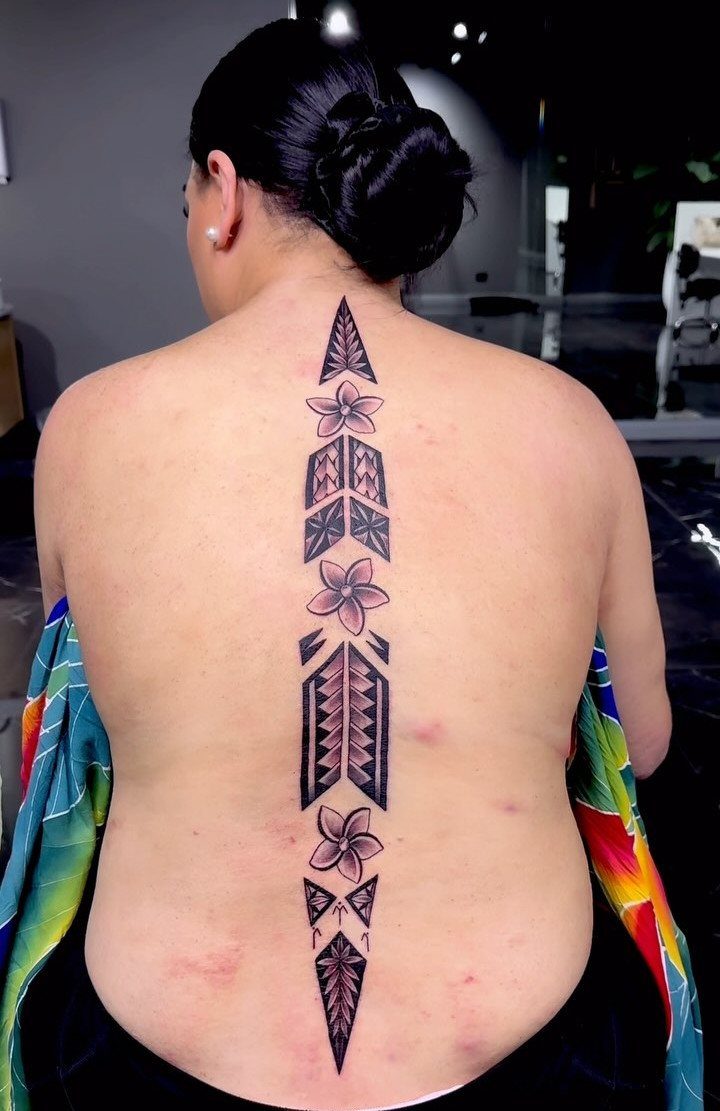 Woman's back with a vertical tribal tattoo and plumeria flowers along the spine.