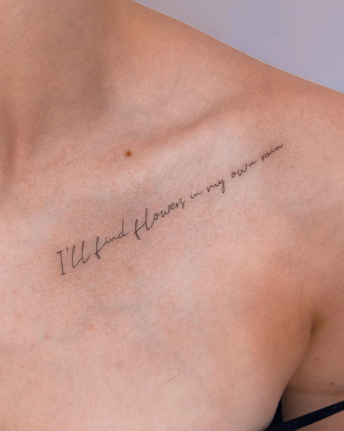 Cursive quote tattoo on a person's collarbone reading I'll find flowers in my own rain