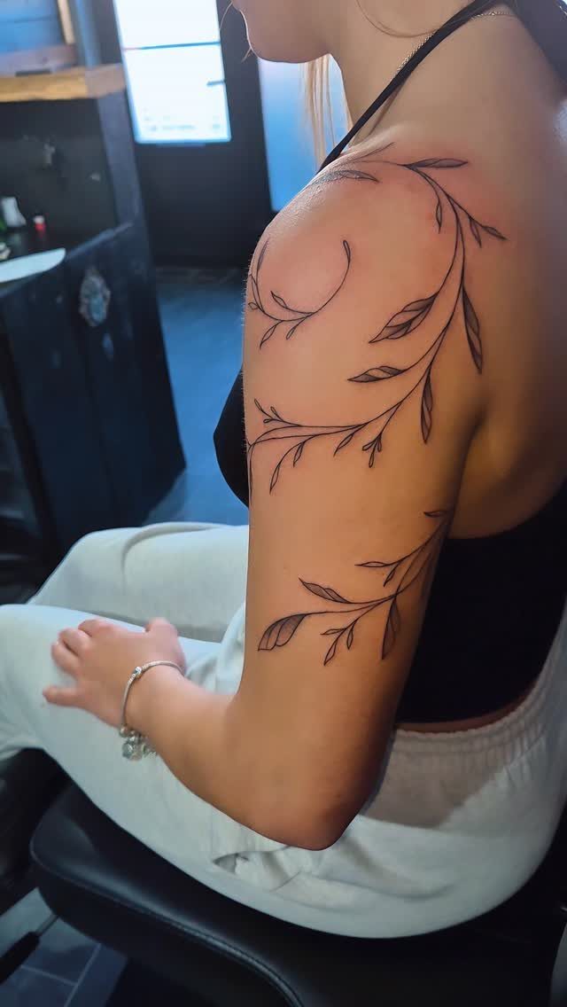 Curving vine tattoo with small leaves on a person's shoulder and upper arm.