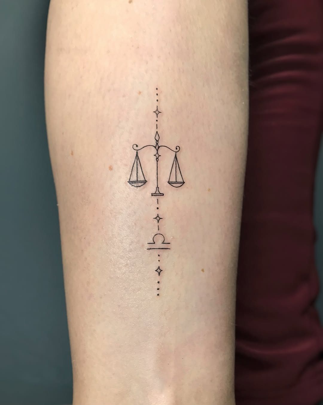 Fine line tattoo of a scale and Libra symbol on a person's arm.